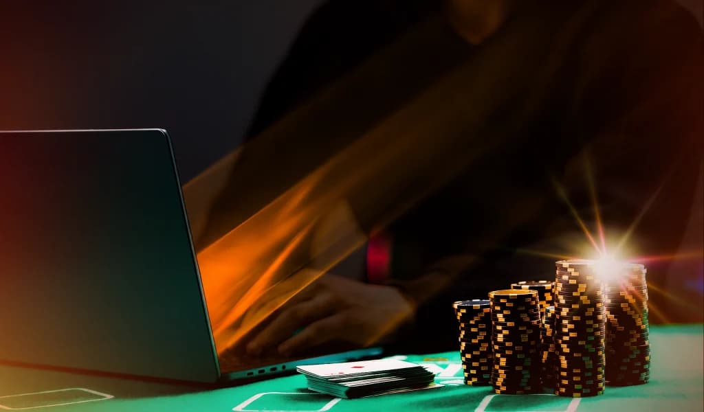 Person playing online poker on a laptop next to stacks of poker chips on a green felt table.
