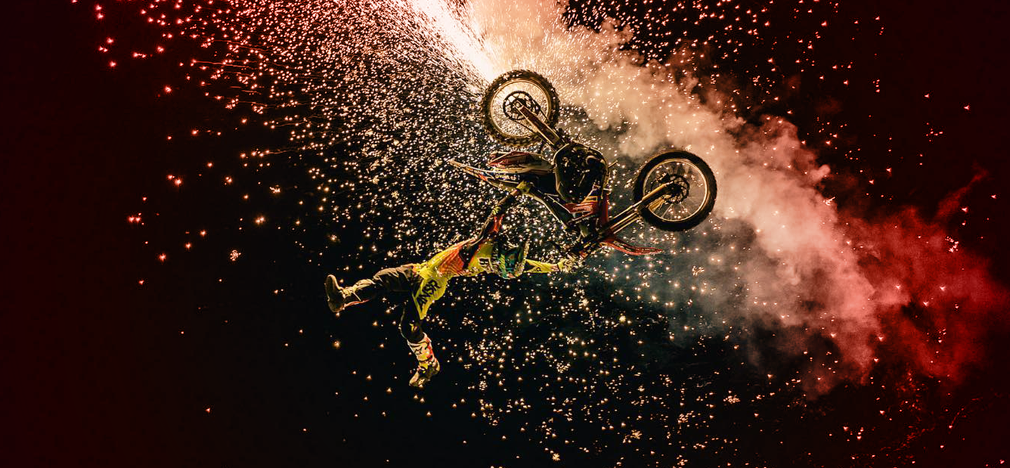 Man doing a flip in the air on a bicycle surrounded with dust and fireworks.