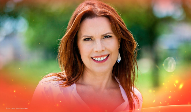 Annie Duke smiling outdoors with auburn hair and white earrings, in front of green foliage and orange lighting.