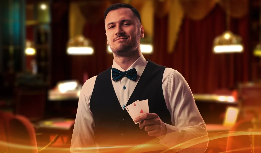 A confident male dealer in a black vest and bow tie holds an ace of hearts in a dimly lit casino with warm lighting.