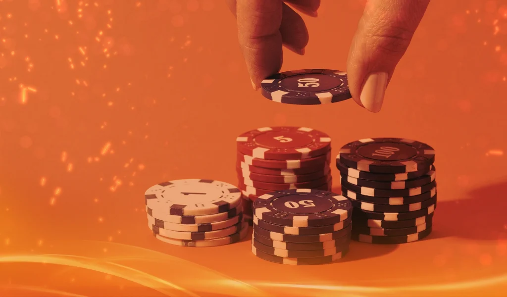 A hand picking up a poker chip from stacks of red, black, and white chips, set against a warm orange background.