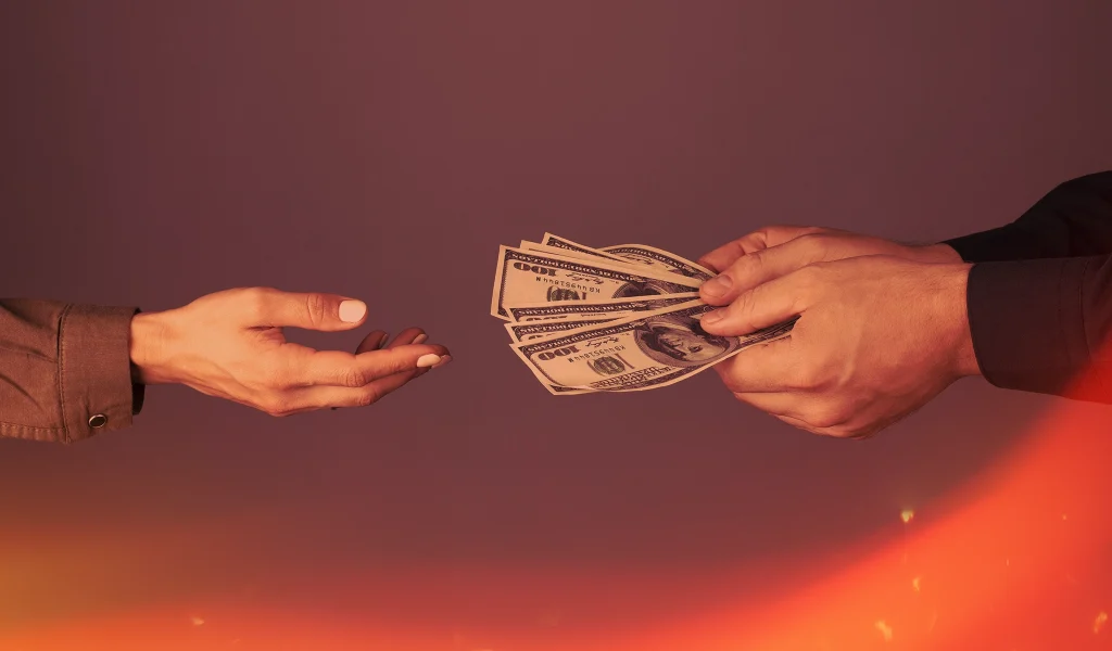Close-up of hands exchanging US dollar bills, with a warm orange glow in the background.