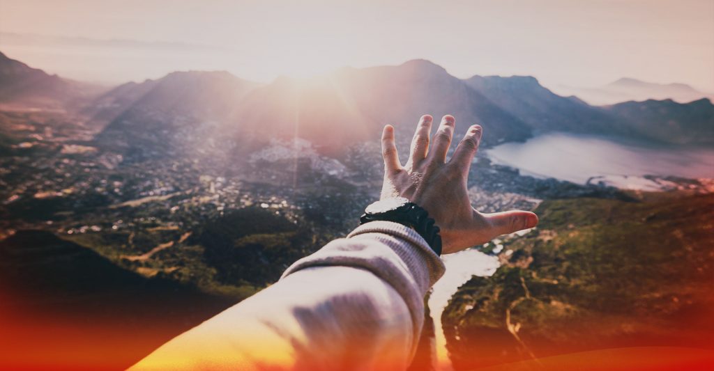 POV of a hand reaching towards the sun over a picturesque mountainous landscape with lakes
